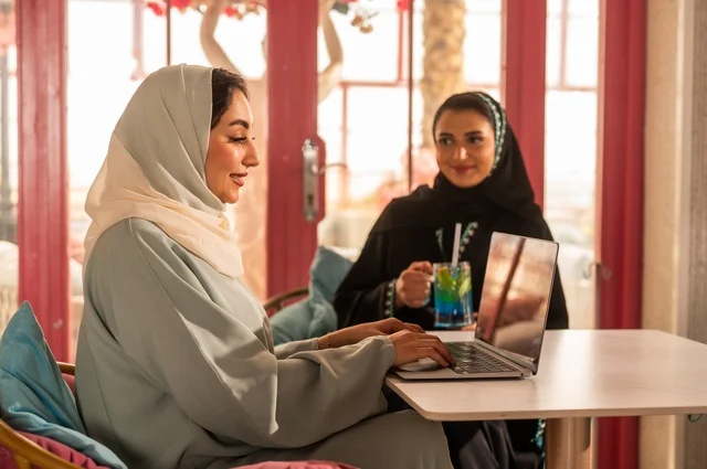 انجاز مهام العمل عن بعد باستخدام الحاسوب المحمول، قضاء أوقات ممتعة مع الأصدقاء، النقاش والحوار في الأجواء الهادئة، مشاركة المشروبات الباردة، صديقتان عربيتان خليجيتان سعوديتان تجلسان في إحدى المقاهي الراقية بإيماءات السعادة والسرور، استخدام جهاز تقني حديث