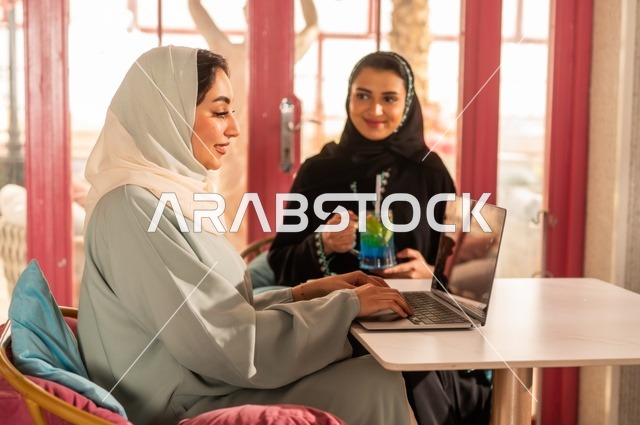 Completing remote work tasks using a laptop, spending enjoyable times with friends, discussing and conversing in a calm atmosphere, sharing cold drinks, two Arab Gulf Saudi friends sitting in a high-end café with gestures of happiness and joy, using modern technology.