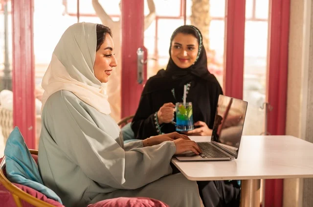 انجاز مهام العمل عن بعد باستخدام الحاسوب المحمول، قضاء أوقات ممتعة مع الأصدقاء، النقاش والحوار في الأجواء الهادئة، مشاركة المشروبات الباردة، صديقتان عربيتان خليجيتان سعوديتان تجلسان في إحدى المقاهي الراقية بإيماءات السعادة والسرور، استخدام جهاز تقني حديث