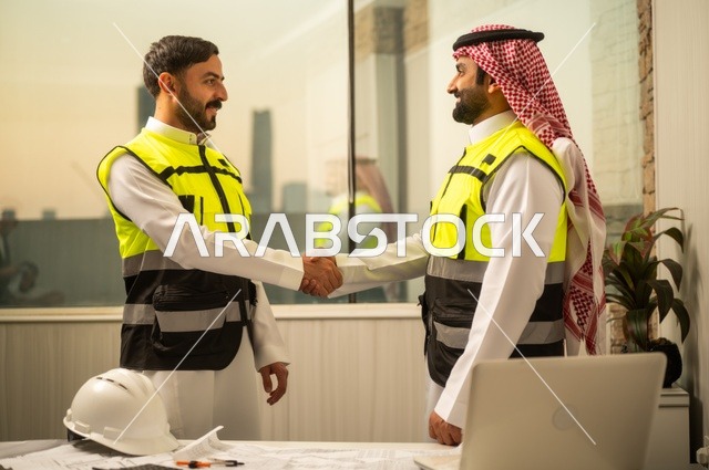 عقد اتفاقيات عمل جديدة ، إيماءات السعادة والسرور، الشركات المختصة بورشات البناء والمقاولات، تصميم وتخطيط المخططات المعمارية، مهندس عربي خليجي سعودي يرتدي الشماغ يقوم بمصافحة مهندس يرتدي الخوذة وسترة الحماية داخل المكتب