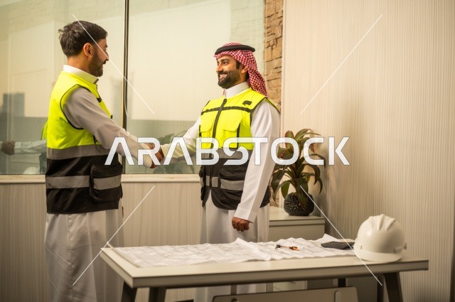 عقد اتفاقيات عمل جديدة ، إيماءات السعادة والسرور، الشركات المختصة بورشات البناء والمقاولات، تصميم وتخطيط المخططات المعمارية، مهندس عربي خليجي سعودي يرتدي الشماغ يقوم بمصافحة مهندس يرتدي الخوذة وسترة الحماية داخل المكتب