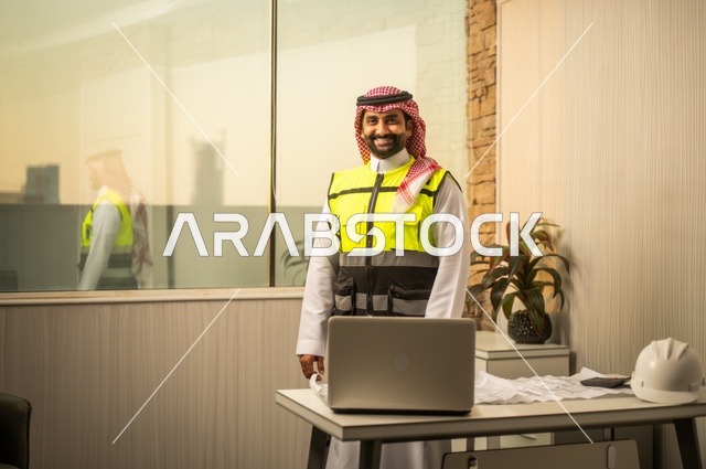 Integrating technology with engineering to complete construction blueprint drawings, auditing and reviewing engineering plans, the concept of engineering and construction, a Saudi Gulf Arab engineer wearing traditional attire, a jacket, and a work helmet sitting at a desk and working on a laptop.