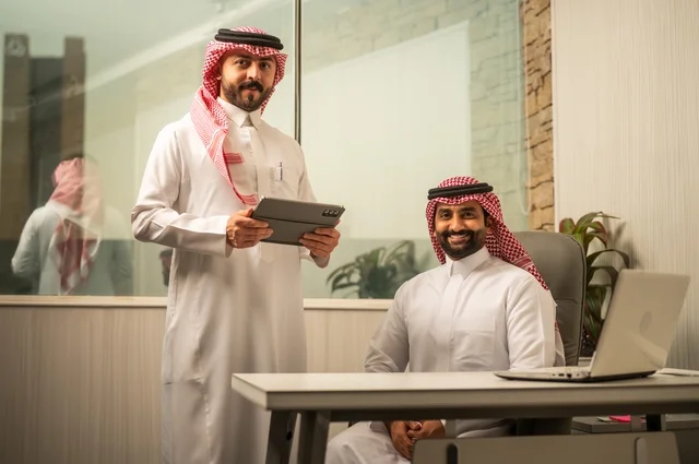 تبادل الخبرات والافكار، دراسة وتحليل المشاريع الجديدة، استخدام جهاز تقني حديث، الوحدة والعمل الجماعي، تطوير العمل وحل المشكلات، رجل عربي خليجي سعودي يرتدي الشماغ والثوب التقليدي يقوم بالعمل برفقة مديره، مهنة ووظيفة سعودية مكتبية