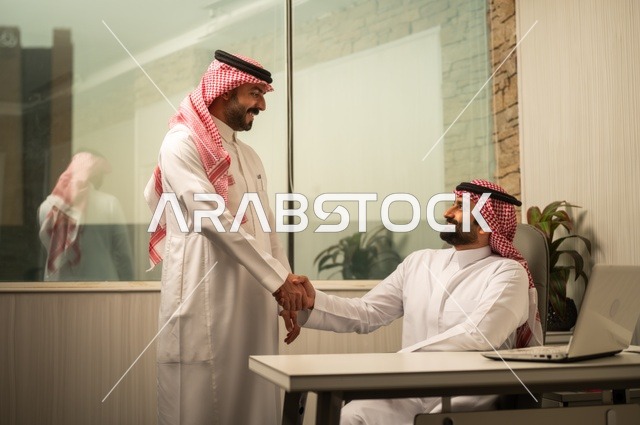 تبادل الخبرات والافكار، دراسة وتحليل المشاريع الجديدة، استخدام جهاز تقني حديث، الوحدة والعمل الجماعي، تطوير العمل وحل المشكلات، رجل عربي خليجي سعودي يرتدي الشماغ والثوب التقليدي يقوم بالعمل برفقة مديره، مهنة ووظيفة سعودية مكتبية