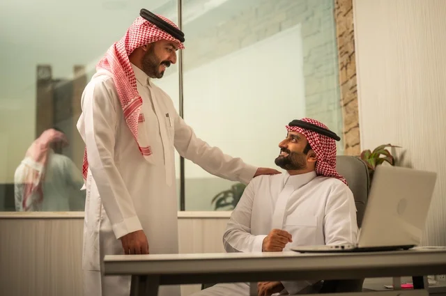 Exchange of experiences and ideas, studying and analyzing new projects, using modern technical devices, unity and teamwork, developing work and solving problems, an Arab Gulf Saudi man wearing the traditional headscarf and thobe working alongside his manager, a Saudi office profession and job.