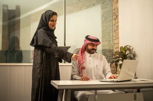 Training employees on new work methods to achieve the company's goals, integrating work with technology and tech, two Arab Gulf Saudi colleagues completing tasks via laptop and tablet, teamwork and the spirit of one team.