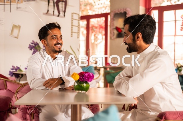 تبادل الأحاديث والحوار بين الأصدقاء، جلسة تسلية وترفيه شبابية، تبادل أطراف الحديث، صداقة وتعارف وبناء علاقات، ايماءات السعادة والسرور، شابان عربيان خليجيان سعوديان يرتديان الثوب الأبيض يجلسان في كوفي شوب راقي، قضاء وقت ممتع في الخارج