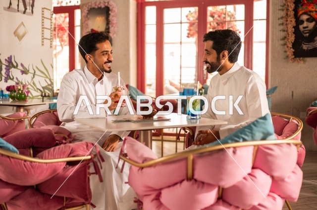 تعابير وجه ابتسامة  ومحبة، تناول المشروبات الباردة، جلسة تسلية وترفيه شبابية، صداقة وتعارف وبناء علاقات، ايماءات السعادة والسرور، شابان عربيان خليجيان سعوديان يرتديان الثوب الأبيض يجلسان في كوفي شوب راقي، قضاء وقت ممتع في الخارج