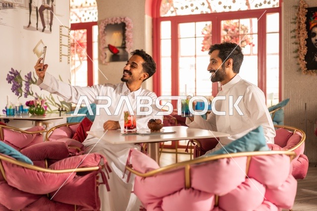 Enjoying cold drinks, taking a memorable selfie for beautiful moments with friends, a session of fun and youthful entertainment, friendship, getting to know each other, and building relationships, gestures of happiness and joy, two young Arab Gulf Saudis wearing white thobes sitting in an upscale coffee shop, having a good time outdoors.
