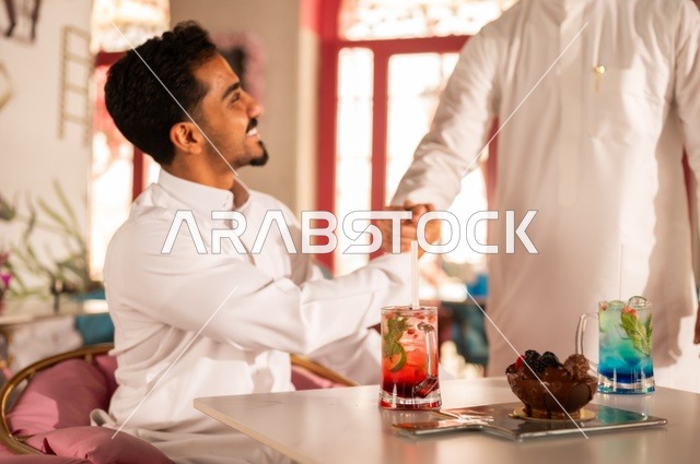 جلسة تسلية وترفيه شبابية، تعابير وجه ويد تدل على المودة والصداقة، تناول المشروبات الباردة،  صداقة وتعارف وبناء علاقات، ايماءات السعادة والسرور، صورة مقربة لشابان عربيان خليجيان سعوديان يرتديان الثوب الأبيض يجلسان في كوفي شوب راقي، قضاء وقت ممتع في الخارج