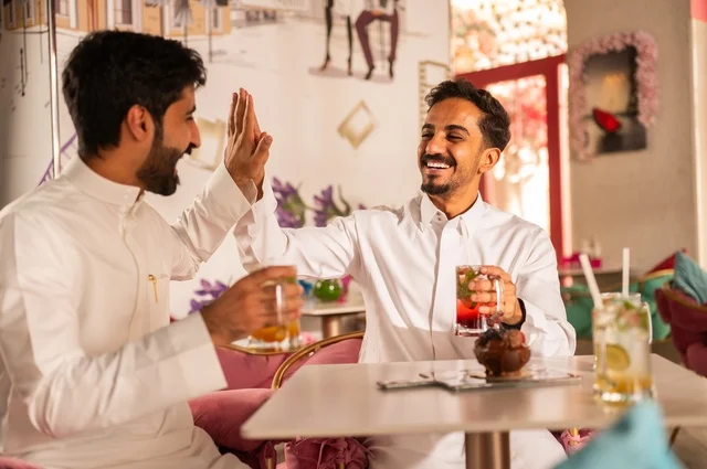 Hi Five Youth is a guide to consensus, a session of youth entertainment and fun, enjoying cold drinks, friendship, getting to know each other, and building relationships, gestures of happiness and joy, two young Arab Gulf Saudis wearing white thobes sitting in an upscale coffee shop, having a good time outdoors.