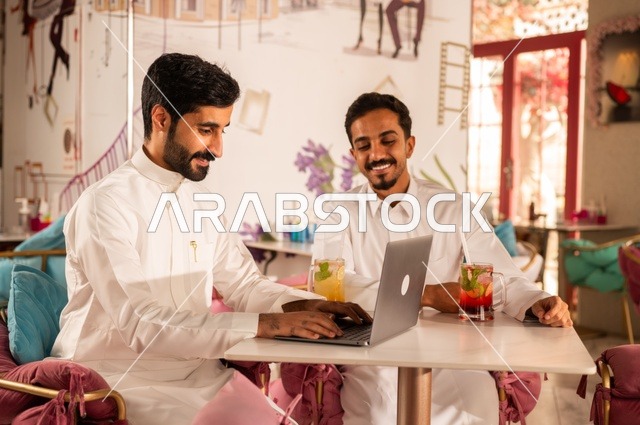 المساعدة في العمل ومشاركات الثقافات بين الأصدقاء، جلسة تسلية وترفيه شبابية، تناول المشروبات الباردة،  صداقة وتعارف وبناء علاقات، ايماءات السعادة والسرور، شابان عربيان خليجيان سعوديان يرتديان الثوب الأبيض يجلسان في كوفي شوب راقي، قضاء وقت ممتع في الخارج