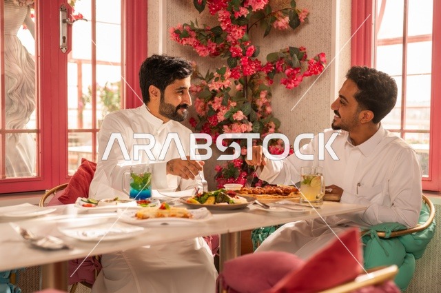 تبادل الأحاديث أثناء تناول الطعام، جلسة تسلية وترفيه شبابية، صداقة وتعارف وبناء علاقات، ايماءات السعادة والسرور، شابان عربيان خليجيان سعوديان يرتديان الثوب الأبيض يجلسان في مطعم راقي أمام طاولة مليئة بالأطعمة الغربية، قضاء وقت ممتع في الخارج