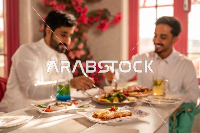 مشاركة الطعام مع الأصدقاء، جلسة تسلية وترفيه شبابية، صداقة وتعارف وبناء علاقات، ايماءات السعادة والسرور، شابان عربيان خليجيان سعوديان يرتديان الثوب الأبيض يجلسان في مطعم راقي أمام طاولة مليئة بالأطعمة الغربية، قضاء وقت ممتع في الخارج