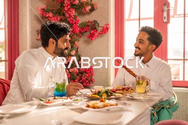 Having cold drinks with lunch, a session of youth entertainment and fun, friendship, getting to know each other, and building relationships, gestures of happiness and joy, two young Arab Gulf Saudis wearing white thobes sitting in an upscale restaurant in front of a table full of Western food, enjoying a good time outdoors.