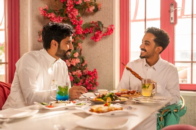 Having cold drinks with lunch, a session of youth entertainment and fun, friendship, getting to know each other, and building relationships, gestures of happiness and joy, two young Arab Gulf Saudis wearing white thobes sitting in an upscale restaurant in front of a table full of Western food, enjoying a good time outdoors.