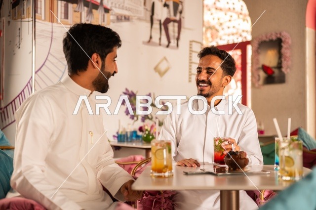 Telling jokes and exchanging funny stories, a session of entertainment and fun for young people, enjoying cold drinks, friendship and getting to know each other and building relationships, gestures of happiness and joy, two young Arab Gulf Saudis wearing white thobes sitting in an upscale coffee shop, spending enjoyable time outdoors.