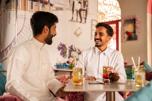 Telling jokes and exchanging funny stories, a session of entertainment and fun for young people, enjoying cold drinks, friendship and getting to know each other and building relationships, gestures of happiness and joy, two young Arab Gulf Saudis wearing white thobes sitting in an upscale coffee shop, spending enjoyable time outdoors.