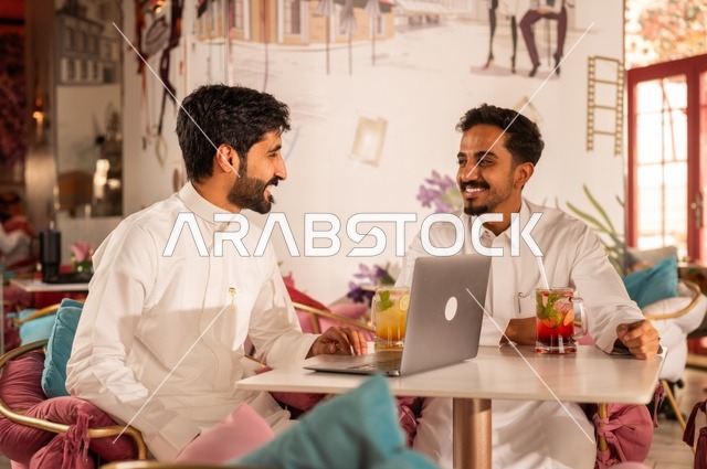 المساعدة في العمل ومشاركات الثقافات بين الأصدقاء، جلسة تسلية وترفيه شبابية، تناول المشروبات الباردة،  صداقة وتعارف وبناء علاقات، ايماءات السعادة والسرور، شابان عربيان خليجيان سعوديان يرتديان الثوب الأبيض يجلسان في كوفي شوب راقي، قضاء وقت ممتع في الخارج