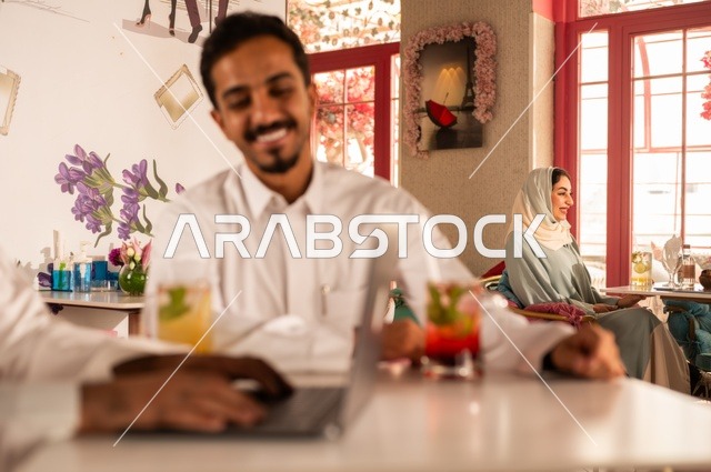The use of technology in daily life, a session of fun and youth entertainment, enjoying cold drinks, friendship and acquaintance and building relationships, gestures of happiness and joy, a close-up image of two young Arab Gulf Saudis wearing white thobes sitting in an upscale coffee shop, spending enjoyable time outdoors.