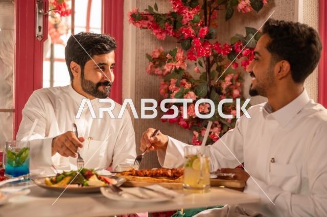 Exchanging conversations while dining, a session of youthful entertainment and fun, friendship and acquaintance, and building relationships, gestures of happiness and joy, two young Arab Gulf Saudis wearing white thobes sitting in an upscale restaurant in front of a table filled with Western foods, enjoying a good time outdoors.