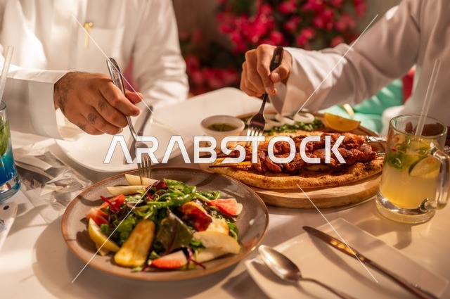 أطباق غربية منوعة، جلسة تسلية وترفيه شبابية، صداقة وتعارف وبناء علاقات، ايماءات السعادة والسرور، صورة مقربة لشابان عربيان خليجيان سعوديان يرتديان الثوب الأبيض يجلسان في مطعم راقي أمام طاولة مليئة بالأطعمة، قضاء وقت ممتع في الخارج