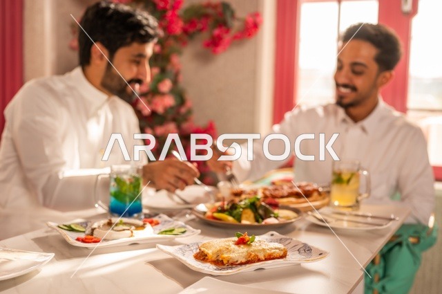 مشاركة الطعام مع الأصدقاء، جلسة تسلية وترفيه شبابية، صداقة وتعارف وبناء علاقات، ايماءات السعادة والسرور، شابان عربيان خليجيان سعوديان يرتديان الثوب الأبيض يجلسان في مطعم راقي أمام طاولة مليئة بالأطعمة الغربية، قضاء وقت ممتع في الخارج