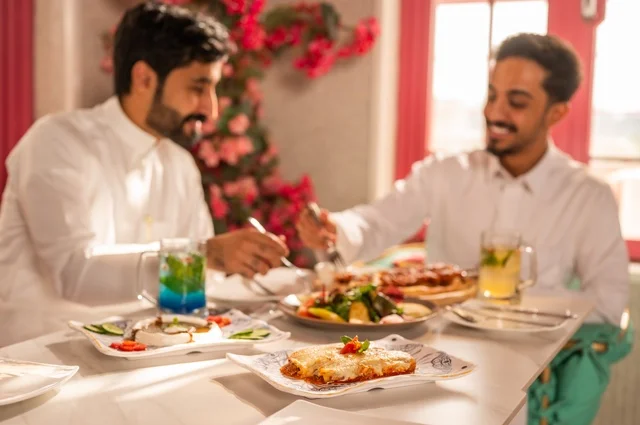 مشاركة الطعام مع الأصدقاء، جلسة تسلية وترفيه شبابية، صداقة وتعارف وبناء علاقات، ايماءات السعادة والسرور، شابان عربيان خليجيان سعوديان يرتديان الثوب الأبيض يجلسان في مطعم راقي أمام طاولة مليئة بالأطعمة الغربية، قضاء وقت ممتع في الخارج