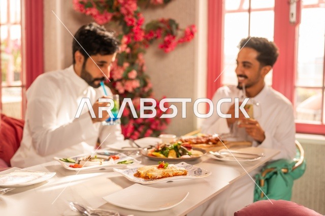 تناول المشروبات الباردة مع وجبة الغداء، جلسة تسلية وترفيه شبابية، صداقة وتعارف وبناء علاقات، ايماءات السعادة والسرور، شابان عربيان خليجيان سعوديان يرتديان الثوب الأبيض يجلسان في مطعم راقي أمام طاولة مليئة بالأطعمة الغربية، قضاء وقت ممتع في الخارج