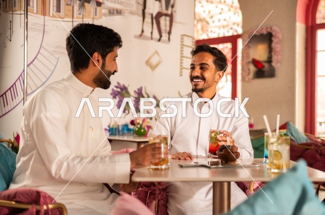 القاء النكت وتبادل القصص الطريفة، جلسة تسلية وترفيه شبابية، تناول المشروبات الباردة،  صداقة وتعارف وبناء علاقات، ايماءات السعادة والسرور، شابان عربيان خليجيان سعوديان يرتديان الثوب الأبيض يجلسان في كوفي شوب راقي، قضاء وقت ممتع في الخارج
