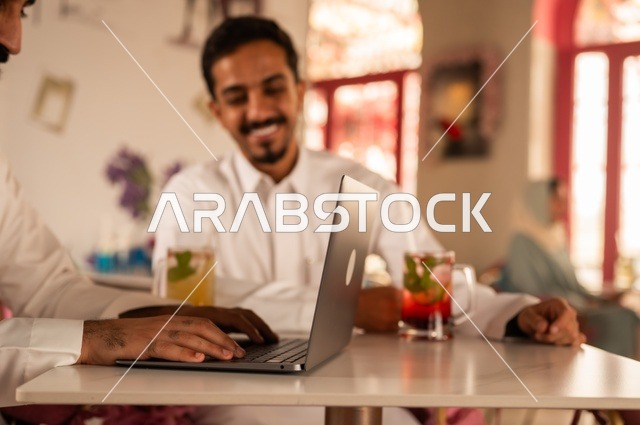 استخدام التكنولوجيا في الحياة اليومية، جلسة تسلية وترفيه شبابية، تناول المشروبات الباردة،  صداقة وتعارف وبناء علاقات، ايماءات السعادة والسرور، صورة مقربة لشابان عربيان خليجيان سعوديان يرتديان الثوب الأبيض يجلسان في كوفي شوب راقي، قضاء وقت ممتع في الخارج