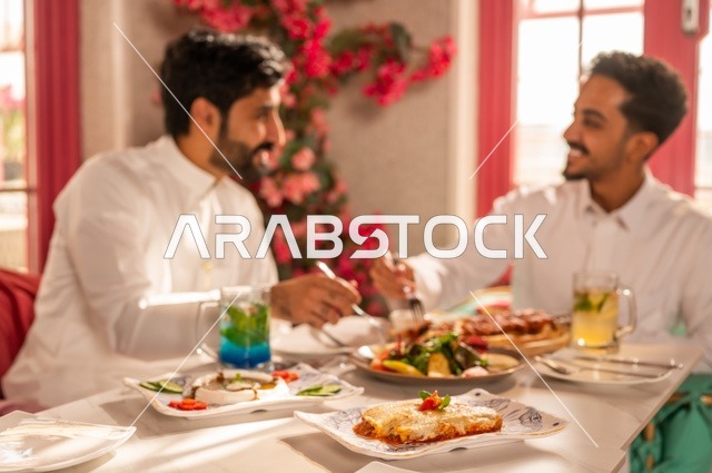 مشاركة الطعام مع الأصدقاء، جلسة تسلية وترفيه شبابية، صداقة وتعارف وبناء علاقات، ايماءات السعادة والسرور، شابان عربيان خليجيان سعوديان يرتديان الثوب الأبيض يجلسان في مطعم راقي أمام طاولة مليئة بالأطعمة الغربية، قضاء وقت ممتع في الخارج