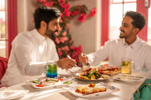Sharing food with friends, a session of youth entertainment and fun, friendship, getting to know each other, and building relationships, gestures of happiness and joy, two young Arab Gulf Saudis wearing white thobes sitting in an upscale restaurant in front of a table full of Western foods, enjoying a good time outdoors.