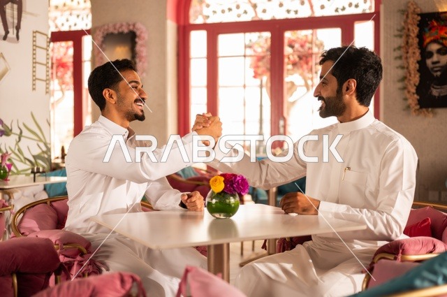 مصافحة شبابية حارة، جلسة تسلية وترفيه شبابية، تبادل أطراف الحديث، صداقة وتعارف وبناء علاقات، ايماءات السعادة والسرور، شابان عربيان خليجيان سعوديان يرتديان الثوب الأبيض يجلسان في كوفي شوب راقي، قضاء وقت ممتع في الخارج، تبادل الأحاديث والحوار بين الأصدقاء