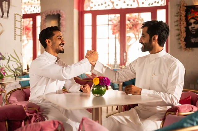 مصافحة شبابية حارة، جلسة تسلية وترفيه شبابية، تبادل أطراف الحديث، صداقة وتعارف وبناء علاقات، ايماءات السعادة والسرور، شابان عربيان خليجيان سعوديان يرتديان الثوب الأبيض يجلسان في كوفي شوب راقي، قضاء وقت ممتع في الخارج، تبادل الأحاديث والحوار بين الأصدقاء