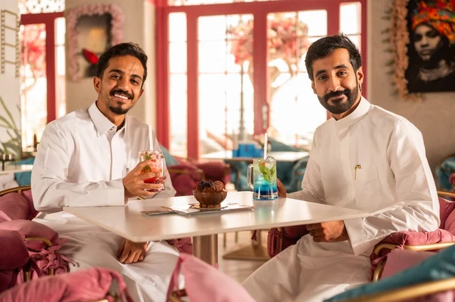 تعابير وجه ابتسامة  ومحبة، تناول المشروبات الباردة، جلسة تسلية وترفيه شبابية، صداقة وتعارف وبناء علاقات، ايماءات السعادة والسرور، شابان عربيان خليجيان سعوديان يرتديان الثوب الأبيض يجلسان في كوفي شوب راقي، قضاء وقت ممتع في الخارج