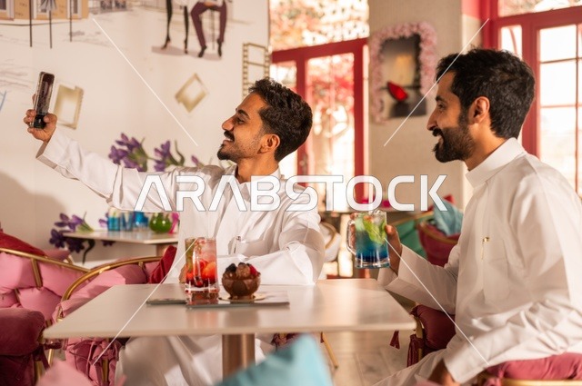 تناول المشروبات الباردة، التقاط سيلفي تذكاري لللحظات الجميلة مع الأصدقاء، جلسة تسلية وترفيه شبابية، صداقة وتعارف وبناء علاقات، ايماءات السعادة والسرور، شابان عربيان خليجيان سعوديان يرتديان الثوب الأبيض يجلسان في كوفي شوب راقي، قضاء وقت ممتع في الخارج