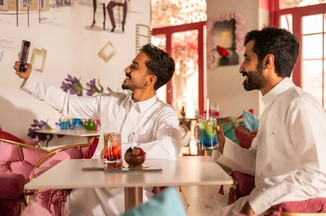 تناول المشروبات الباردة، التقاط سيلفي تذكاري لللحظات الجميلة مع الأصدقاء، جلسة تسلية وترفيه شبابية، صداقة وتعارف وبناء علاقات، ايماءات السعادة والسرور، شابان عربيان خليجيان سعوديان يرتديان الثوب الأبيض يجلسان في كوفي شوب راقي، قضاء وقت ممتع في الخارج