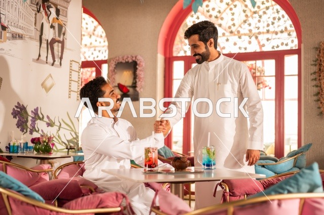 مصافحة وسلام تدل على المودة والصداقة، جلسة تسلية وترفيه شبابية، تناول المشروبات الباردة،  صداقة وتعارف وبناء علاقات، ايماءات السعادة والسرور، صورة مقربة لشابان عربيان خليجيان سعوديان يرتديان الثوب الأبيض يجلسان في كوفي شوب راقي، قضاء وقت ممتع في الخارج