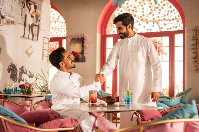 A handshake and greeting indicate affection and friendship, a session of entertainment and youth fun, enjoying cold drinks, friendship and acquaintance and building relationships, gestures of happiness and joy, a close-up image of two young Arab Gulf Saudis wearing white thobes sitting in an upscale coffee shop, spending enjoyable time outdoors.