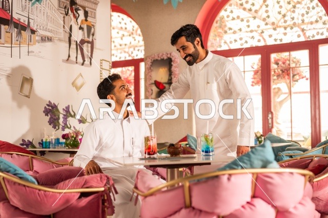 تعابير وجه ويد تدل على المودة والصداقة، تناول المشروبات الباردة، جلسة تسلية وترفيه شبابية، صداقة وتعارف وبناء علاقات، ايماءات السعادة والسرور، شابان عربيان خليجيان سعوديان يرتديان الثوب الأبيض يجلسان في كوفي شوب راقي، قضاء وقت ممتع في الخارج