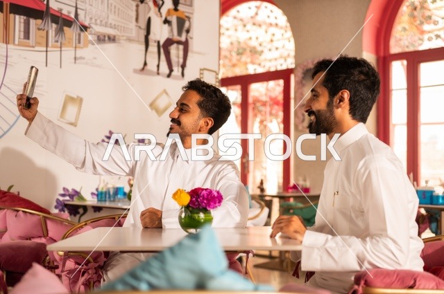 Taking a commemorative selfie for beautiful moments with friends, a session of youth entertainment and fun, exchanging conversations, friendship and acquaintance, building relationships, gestures of happiness and joy, two young Arab Gulf Saudis wearing white thobes sitting in an upscale coffee shop, enjoying time outdoors.