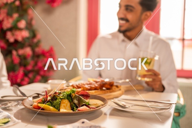 تناول المشروبات الباردة مع وجبة الغداء، جلسة تسلية وترفيه شبابية، صداقة وتعارف وبناء علاقات، ايماءات السعادة والسرور، صورة مقربة لشابان عربيان خليجيان سعوديان يرتديان الثوب الأبيض يجلسان في مطعم راقي أمام طاولة مليئة بالأطعمة الغربية، قضاء وقت ممتع في الخارج