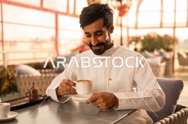 قضاء وقت الفراغ باحتساء المشروب المفضل، تناول المشروبات الشتوية الدافئة، الاستمتاع بالمشروبات الساخنة، رجل عربي خليجي سعودي يرتدي الثوب الأبيض يجلس في كوفي شوب راقي يقوم بشرب القهوة، إيماءات السعادة والسرور