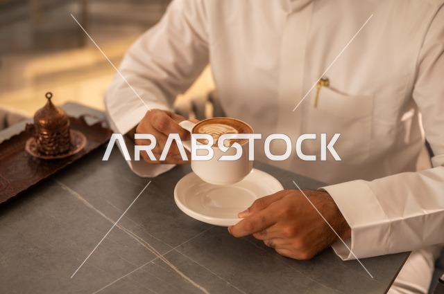 تناول المشروبات الشتوية الدافئة، قضاء وقت الفراغ باحتساء المشروب المفضل،  الاستمتاع بالمشروبات الساخنة،صورة مقربة لرجل عربي خليجي سعودي يرتدي الثوب الأبيض يجلس في كوفي شوب راقي يحمل كوب من القهوة، إيماءات السعادة والسرور