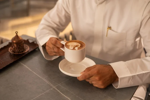 Enjoying warm winter drinks, spending leisure time sipping a favorite beverage, enjoying hot drinks, a close-up of a Saudi Arabian Gulf man wearing a white thobe sitting in a fancy coffee shop holding a cup of coffee, gestures of happiness and joy.