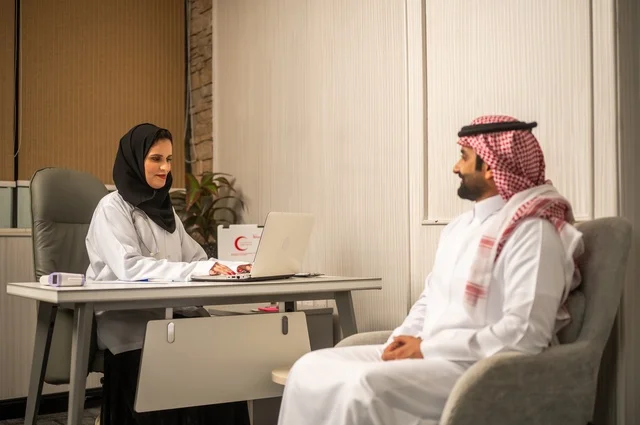 تقديم الاستشارات في المراكز الطبيه, العمل في القطاعات الصحية، متابعة السجل الطبي للمراجعين, تسجيل حالات المرضى على جهاز الحاسوب, طبيبة عربية خليجية سعودية ترتدي المعطف الأبيض تقوم بشرح الحالة المرضية لمريض يرتدي الشماغ والثوب التقليدي، الحوار مع المرضى لكشف الحالة