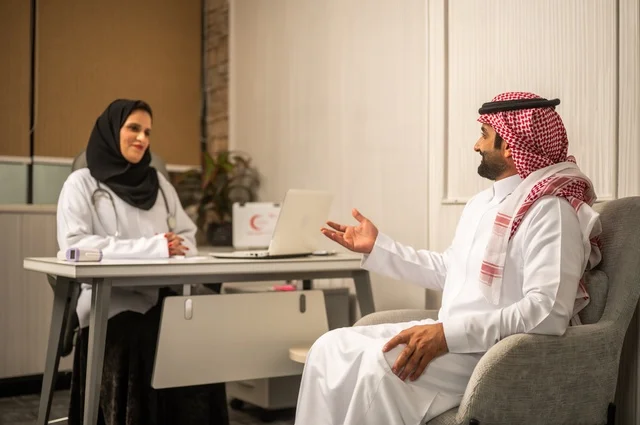 تقديم الاستشارات في المراكز الطبيه, العمل في القطاعات الصحية، متابعة السجل الطبي للمراجعين, تسجيل حالات المرضى على جهاز الحاسوب, طبيبة عربية خليجية سعودية ترتدي المعطف الأبيض تقوم بشرح الحالة المرضية لمريض يرتدي الشماغ والثوب التقليدي، الحوار مع المرضى لكشف الحالة