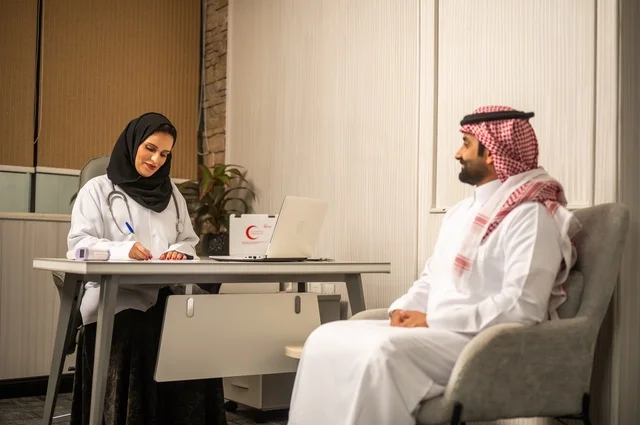 تقديم الاستشارات في المراكز الطبيه, العمل في القطاعات الصحية، متابعة السجل الطبي للمراجعين, تسجيل حالات المرضى على جهاز الحاسوب, طبيبة عربية خليجية سعودية ترتدي المعطف الأبيض تقوم بشرح الحالة المرضية لمريض يرتدي الشماغ والثوب التقليدي، الحوار مع المرضى لكشف الحالة