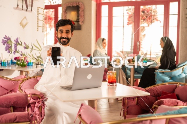 A black background displayed on a mobile phone in an upscale coffee shop, spending leisure time sipping a favorite drink, enjoying cold beverages, a picture of a Saudi Arabian Gulf man wearing a white thobe sitting in a café at a laptop table, gestures of happiness and joy, with Saudi women sitting in the background.
